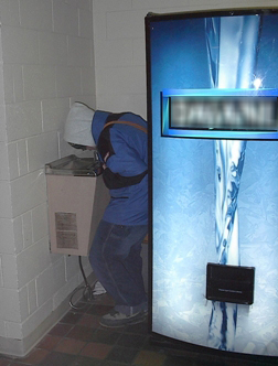 Young man sqeezing past a water bottle machine to access a cramped water fountain